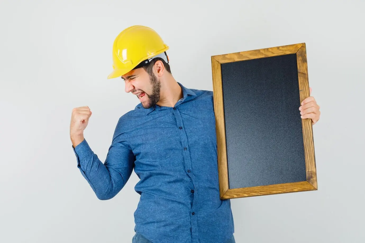 Junger Mann im Hemd, mit Helm, hält eine schwarze Tafel, zeigt eine Siegergeste und blickt glückselig, Vorderansicht.